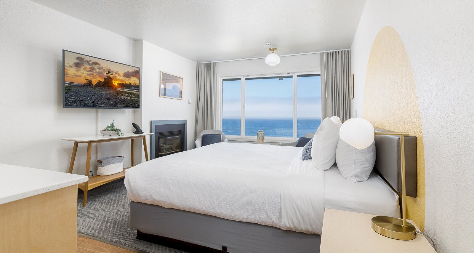 A modern hotel bedroom with a large bed, ocean view through windows, a wall TV, fireplace, and neutral decor.
