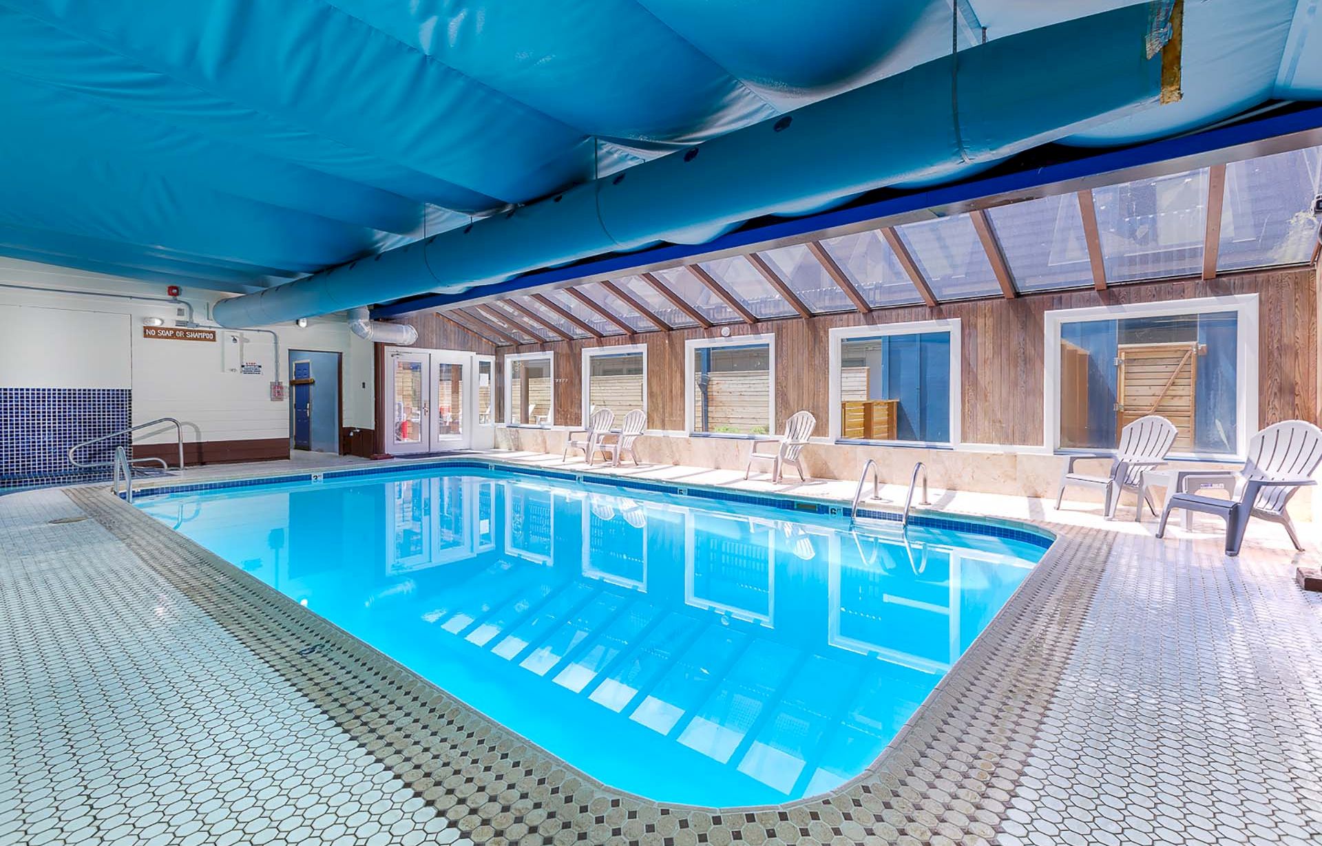 Indoor swimming pool with a blue cover, mosaic deck, and rows of patio chairs in a bright indoor wooden hall.