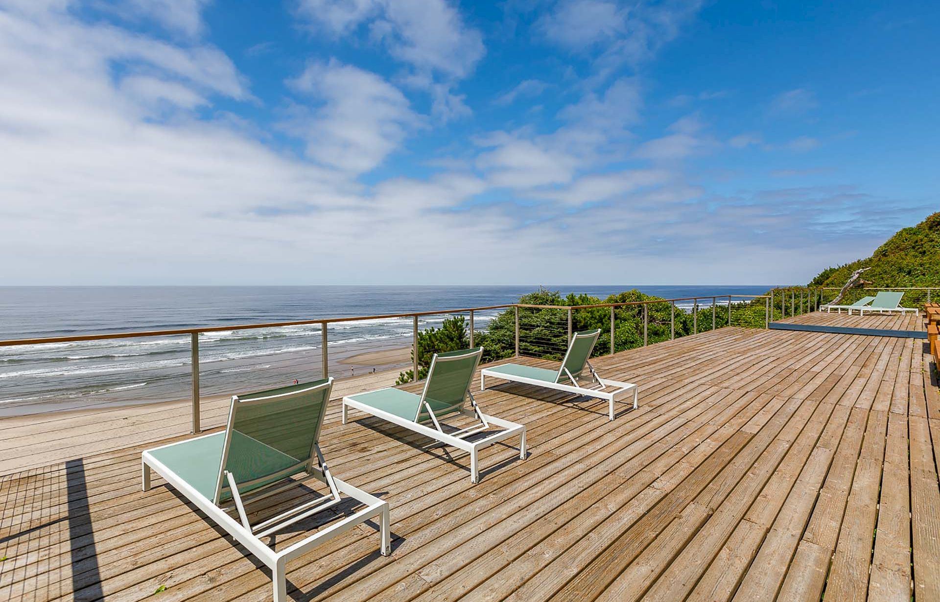 A seaside wooden deck with green lounge chairs and railings, overlooking a sandy beach, rolling waves, and a blue sky with clouds.