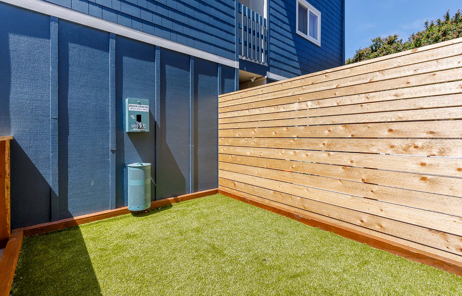 A backyard with artificial grass, a blue wall and fence, a wooden privacy panel, and a small outdoor trash can.