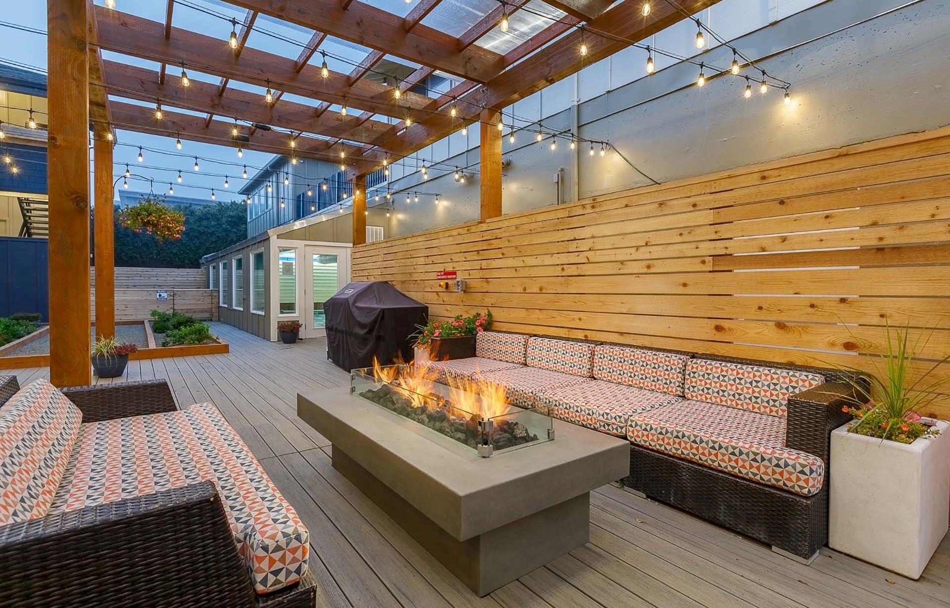 A cozy outdoor patio with wooden pergola, string lights, cushioned seating, and a gas fire pit for warmth and ambiance.