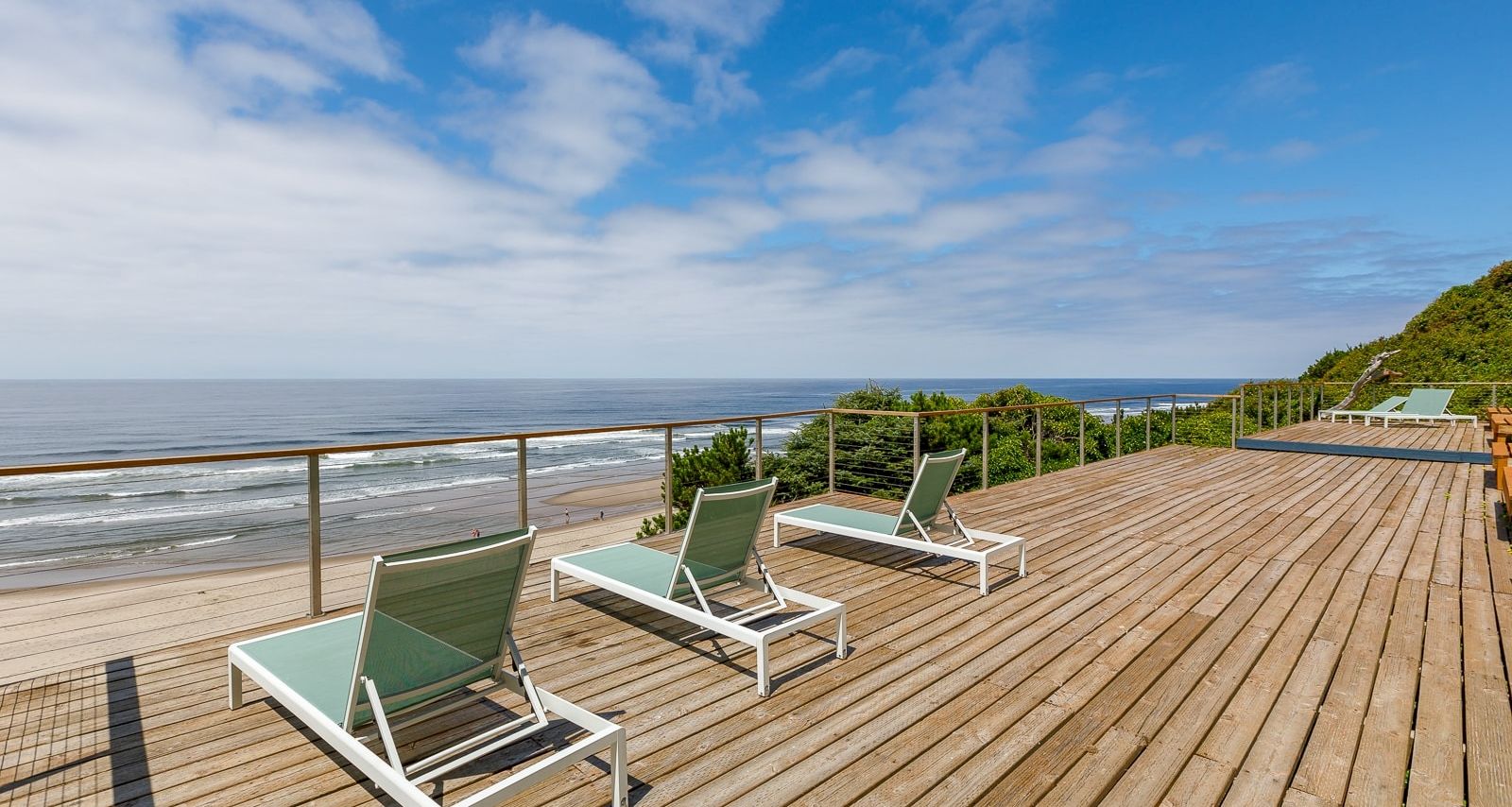 A wooden oceanfront deck with two green lounge chairs, a railing, and waves under a bright blue sky with scattered clouds.