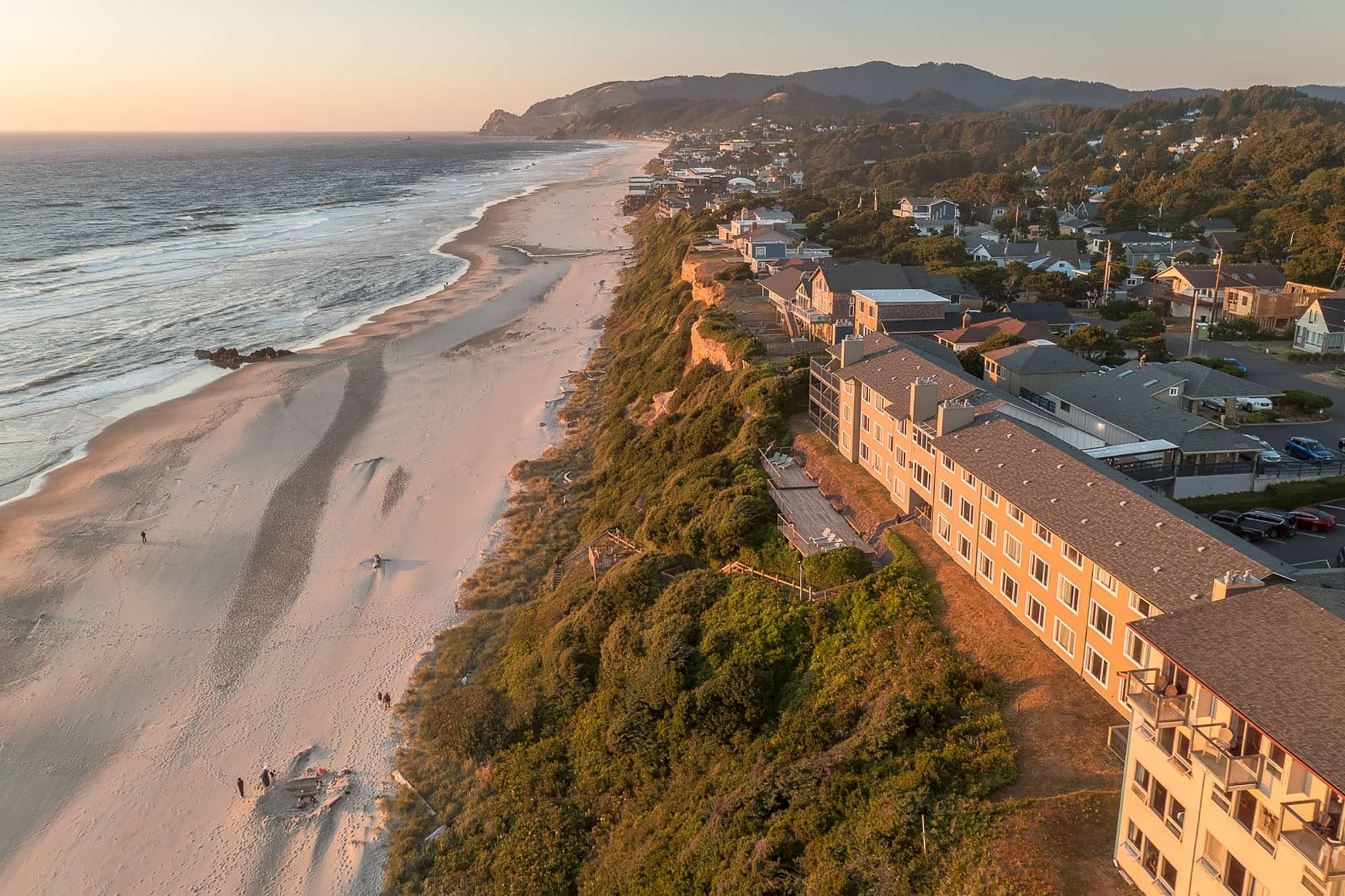 Aerial view of a coastal town at sunset, with a sandy beach, waves, and hillside houses and hotels along the shore.