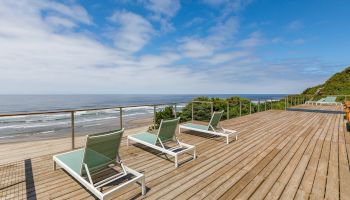 Oceanfront wooden deck with railing and three green lounge chairs, overlooking the beach and choppy waves under a blue sky.
