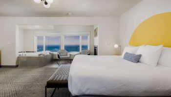 A modern hotel room with a large bed, seaside view windows, lounge chairs, a ceiling lamp, and a yellow wall accent.