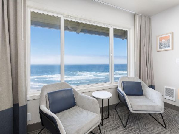 Cozy seaside room with two lounge chairs and small round table facing a large ocean view through bright windows.