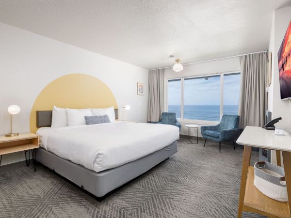 A modern hotel room with a large bed, seating by a window showing the ocean, and a wall-mounted TV.