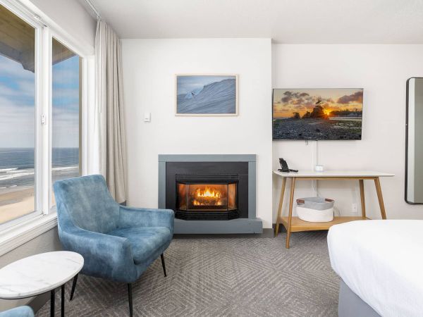 A bright coastal living room with ocean view, blue armchair, lit fireplace, wall art, desk, and a standing mirror.