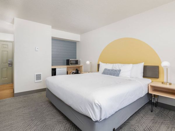 A modern bedroom with a neatly made bed, yellow arched headboard, bedside lamp, and a microwave on a small counter.
