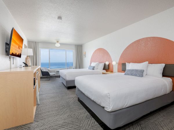 A modern hotel bedroom with twin beds, ocean view through large windows, grey curtains, and a wall TV.