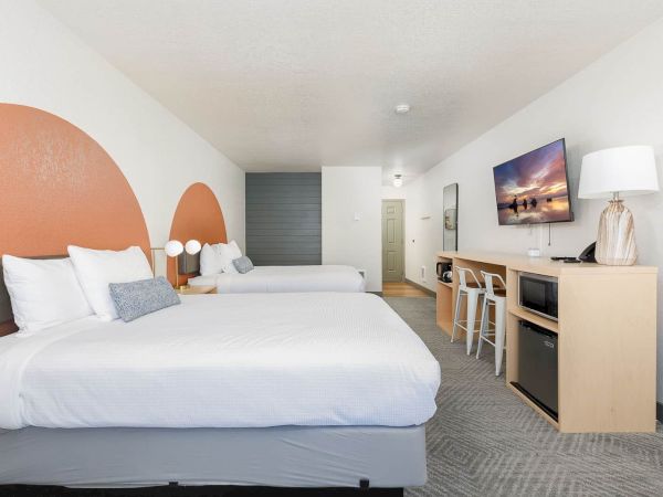 A modern hotel room with twin beds, a desk, TV, lamps, and neutral decor.