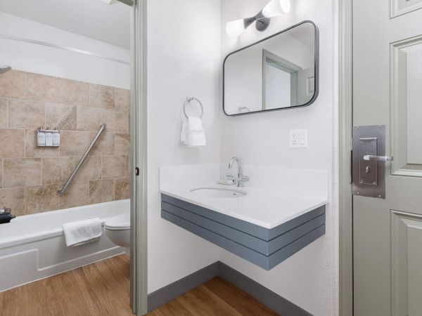 Bright bathroom with a tiled shower, accessible grab bar, gray vanity and sink, illuminated round mirror, towels, and toilet.