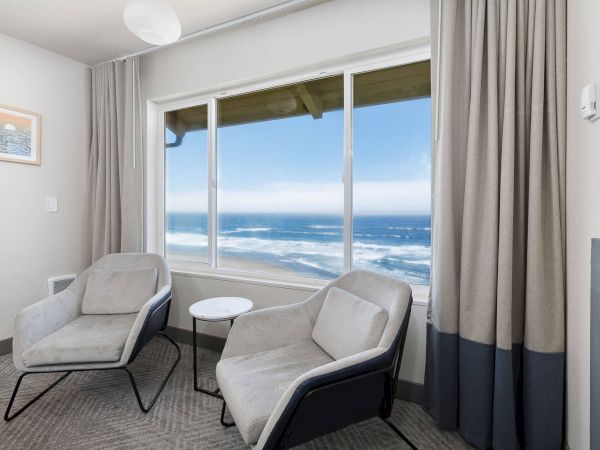 Bright lounge by ocean-view windows with two gray chairs, a small white table, and floor-to-ceiling curtains beside a fireplace.