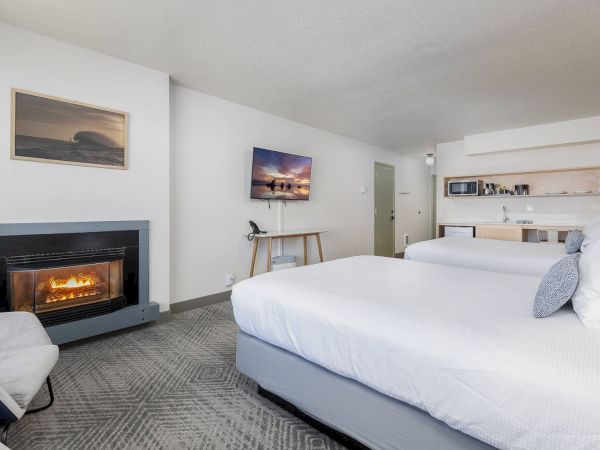 A bright hotel room with two beds, a lit fireplace, wall TVs, and a small kitchenette with coffee supplies.