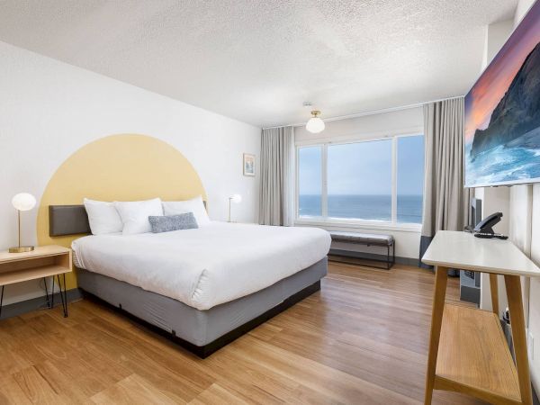 A modern bedroom with a large bed, bedside tables, curtains, ocean-view window, and a TV with desk by the wall.