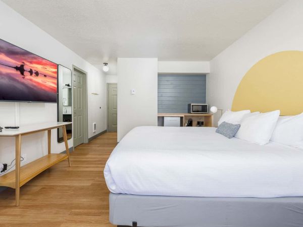 A modern hotel bedroom: neatly made white bed, wall TV, wooden desk, and a microwave on a cabinet.