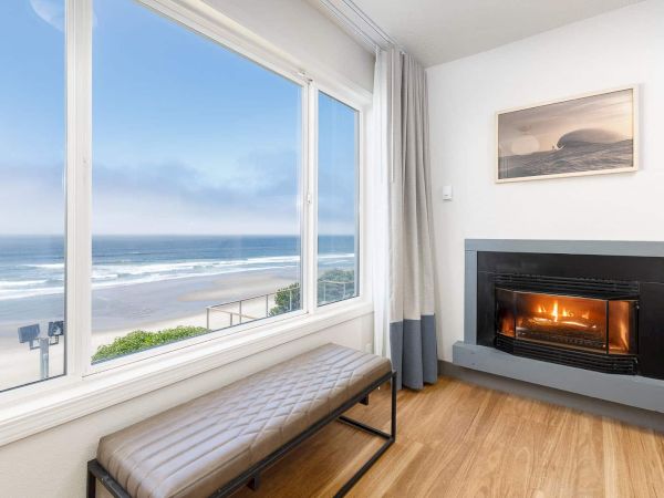 A bright seaside room with a large window view, a cushioned bench, and a crackling fireplace by the wall.