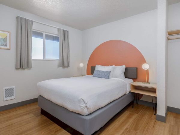 A bright bedroom with a gray bed, white linens, orange headboard, nightstand, window with curtains, and a floor heater.