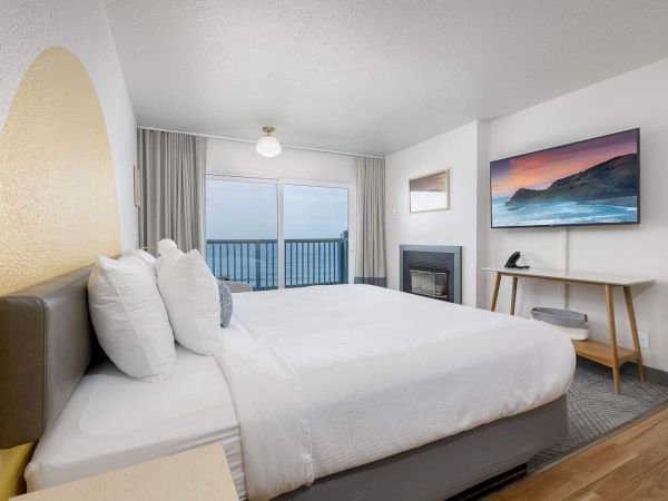 A bright modern bedroom with a large white bed, ocean-view balcony, curtains, fireplace, and framed wall art.