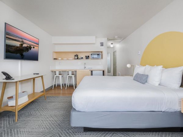 A bright hotel room with a large bed, desk and wall TV, plus a small kitchenette in the back.