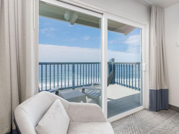 A bright living room with beige curtains and sofa, opening onto a balcony with ocean views and white railings.