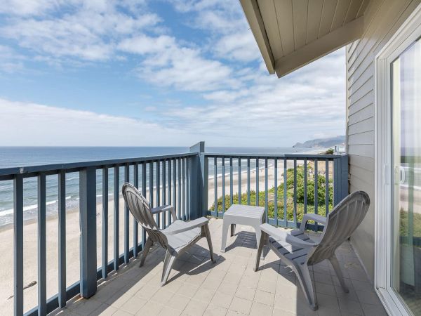 A seaside balcony with two white chairs and a table, overlooking the ocean and sandy beach under a partly cloudy sky.