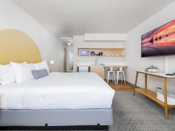 A modern studio room with a large bed, small kitchenette, bar stools, and a wall-mounted sunset TV.