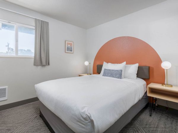 A tidy hotel bedroom with a white bed, orange arched headboard, two lamps, window, and framed wall art.