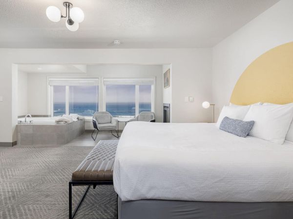 A bright modern hotel bedroom with a large bed, ocean-view windows, and a cozy seating area, lit by a ceiling chandelier.