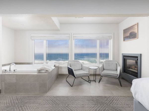 A bright seaside lounge with a soaking tub, two gray chairs, a small table, and an ocean view by a fireplace.