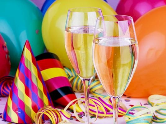 A festive birthday scene with colorful balloons, party hats, and two glasses of champagne on a table.
