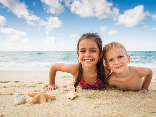 Two children smile in the sand at a sunny beach, with ocean waves and a blue sky behind them.