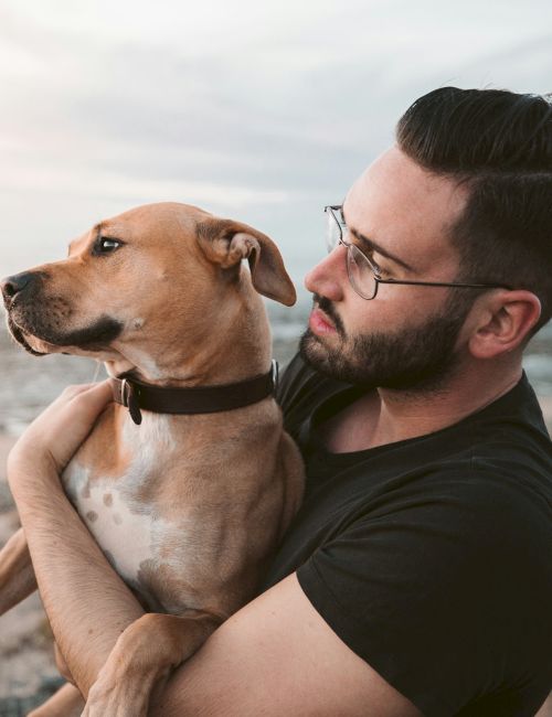 A man holds a brown dog close on a windy beach, sharing a sweet moment by the water.