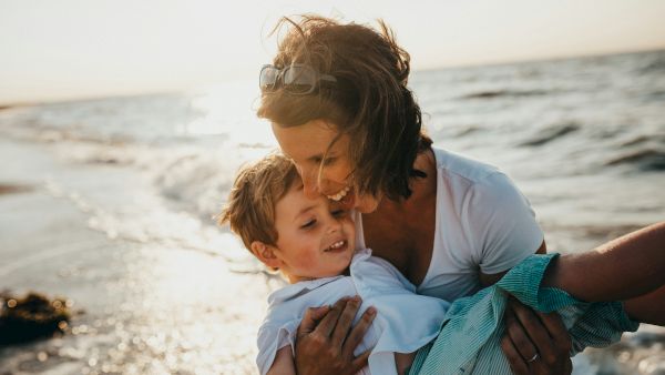 A woman cuddles a smiling child on a sunny beach, with waves and sea sparkle in the background.