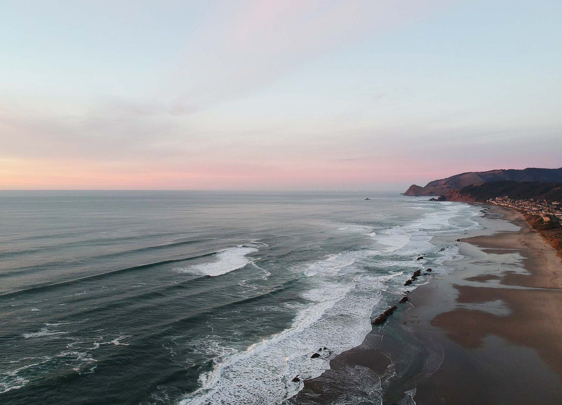 A serene coastal shoreline with waves, a sandy beach, distant cliffs, and a pastel sunset sky over the ocean.