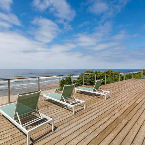 A wooden oceanfront deck with green lounge chairs and a railing overlooking calm waves under a bright blue sky.