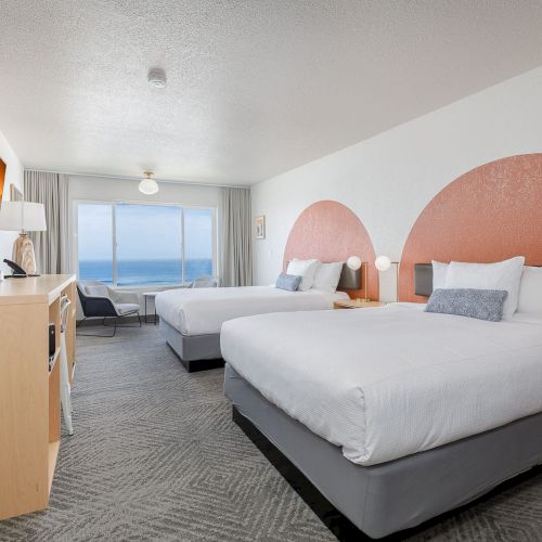 A bright hotel room with two beds, gray curtains, and a wide ocean view through large windows.
