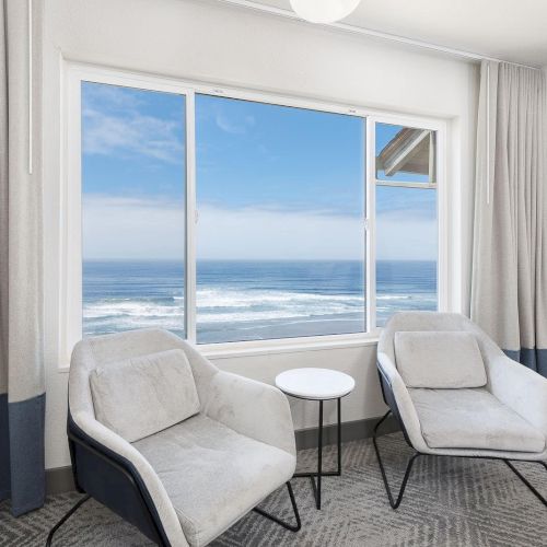 A bright lounge with two beige chairs and a small white table, looking out at ocean waves through big windows.