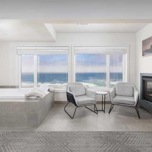 Bright modern living area with ocean-view windows, two grey chairs, small table, a fireplace, and a tiled soaking tub.