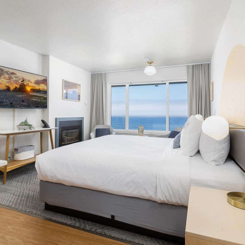Bright hotel bedroom: large white bed, bedside tables, fireplace, and curtains framing an ocean view.
