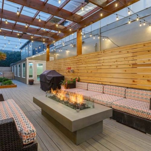 A cozy outdoor patio with a wooden pergola, string lights, cushioned seating, and a central fire pit on a tiled deck.