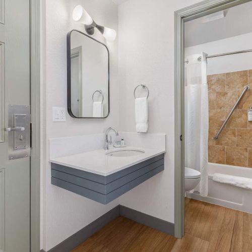A modern bathroom with a vanity sink, wall mirror, toilet, and a tiled shower/tub with white towels.