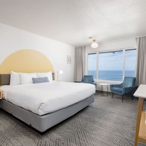 A modern hotel bedroom with a large bed, chairs, TV, and a window showing the ocean view.
