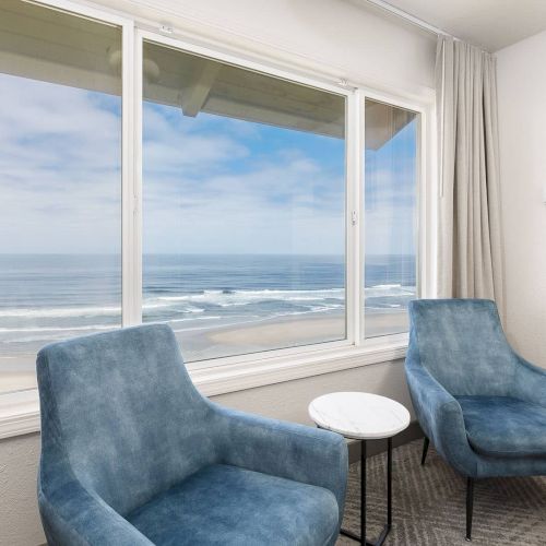 A bright beach-view living room with two blue armchairs and a small white table by large windows, ending in calm sea waves.