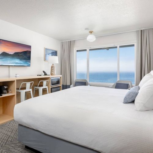 A bright hotel-style bedroom with a large bed, desk/coffee station, ocean view window, and neutral curtains around it.