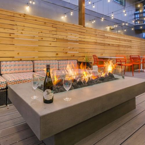 Cozy patio fire pit with flames, orange chairs, patterned cushions, wine bottle and glasses, and string lights overhead.