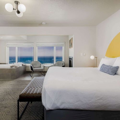 A modern hotel room with a large bed, window ocean view, seating chairs, vanity sink, and globe lights.