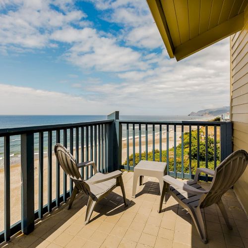 Balcony with patio chairs and table overlooking a sandy beach and ocean under a blue, partly cloudy sky.