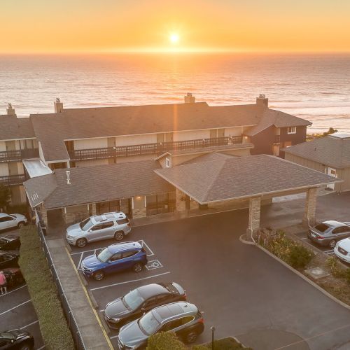 Oceanfront hotel with parked cars, viewed from above at sunrise over calm waves, creating a warm golden seascape.
