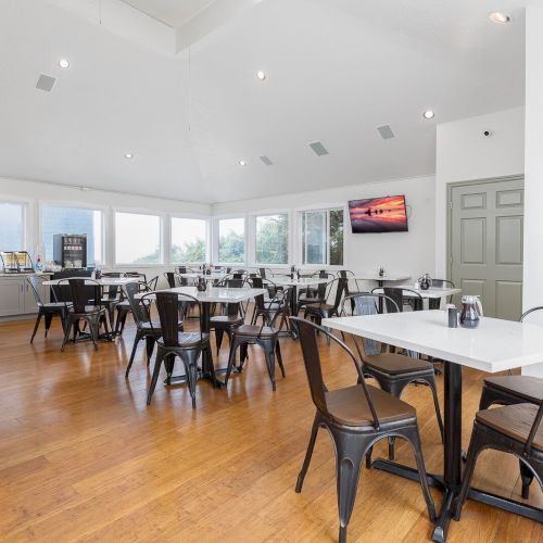 A bright dining hall with rows of metal chairs and white tables, a coffee station on the left, and windows along the back wall.